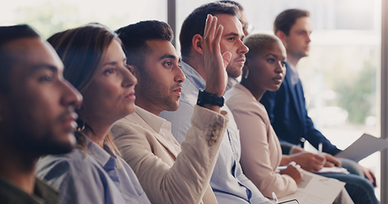 Image of classroom of adult learners.  All are in rapt attention, while one man is raising his hand to ask a question from an unseen instructor